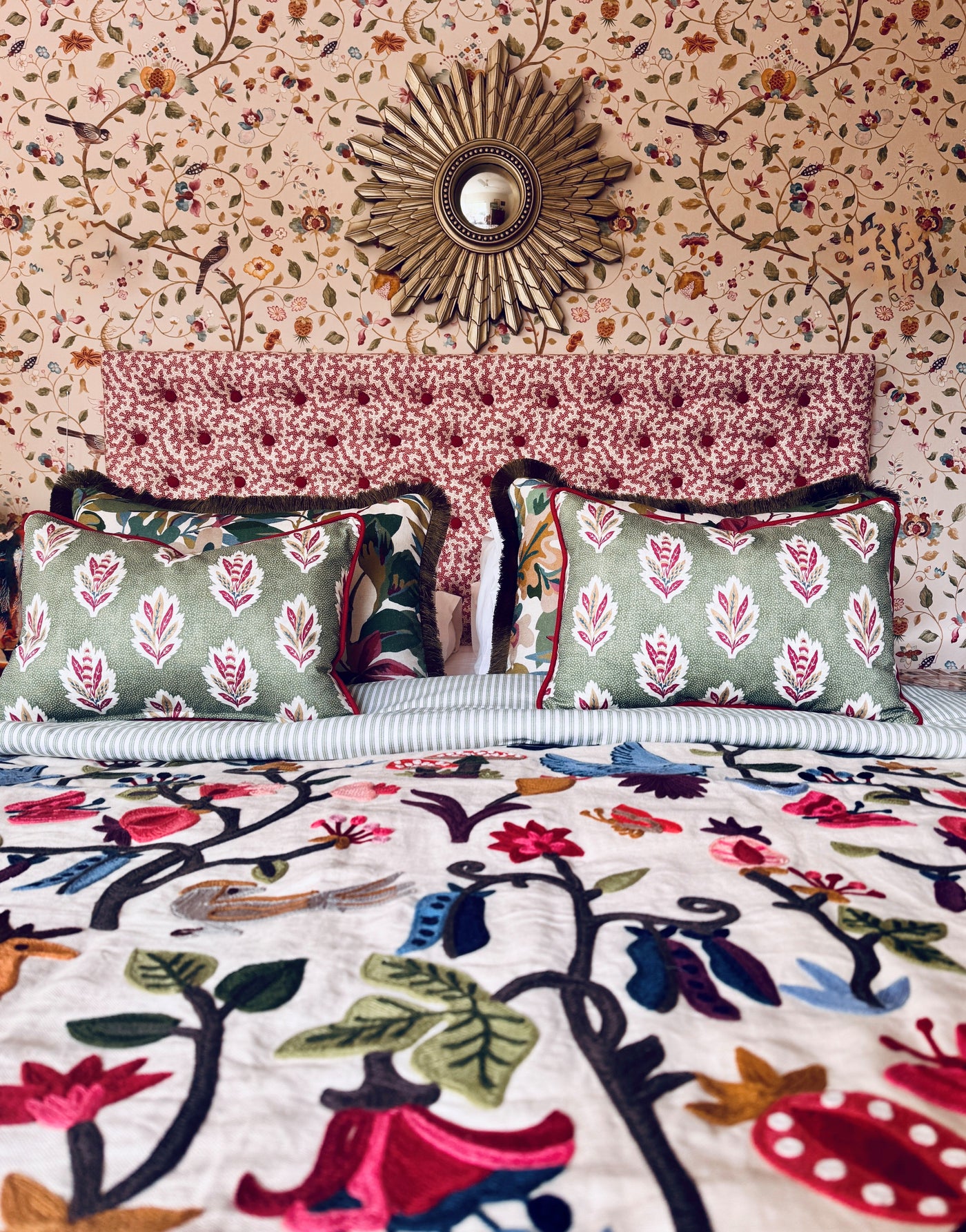 Floral wallpaper behind a patterned upholstered bedhead, green cushions and embroidered bedding.