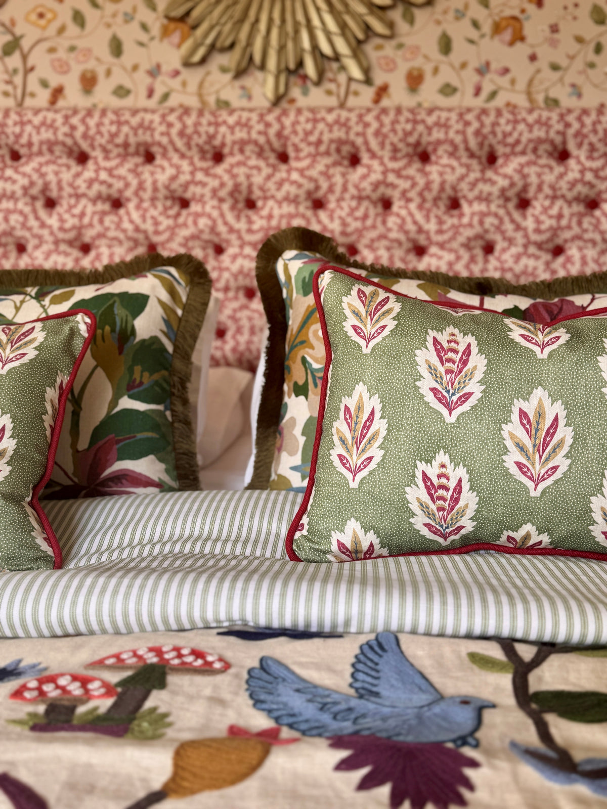 Red-and-cream upholstered bedhead behind patterned green cushions and striped bedding, with botanical wallpaper above.