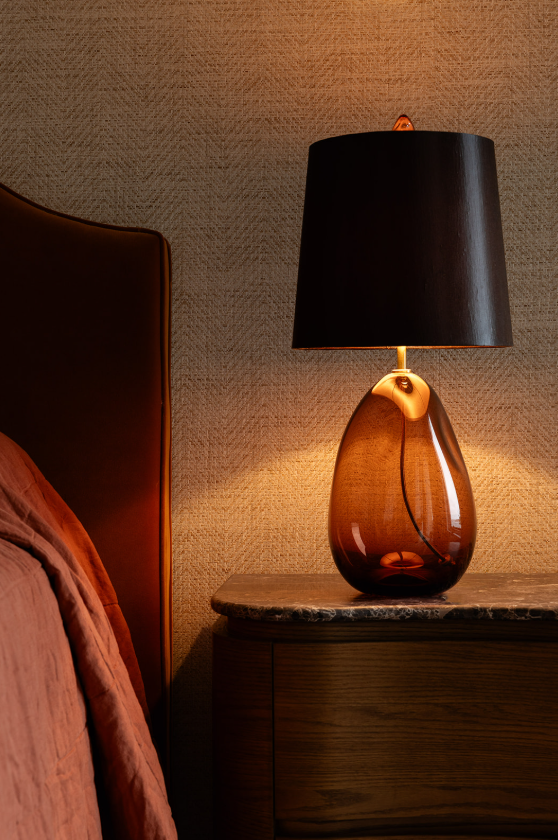 Amber-coloured table lamp with a sculptural base and black shade beside an upholstered bedhead