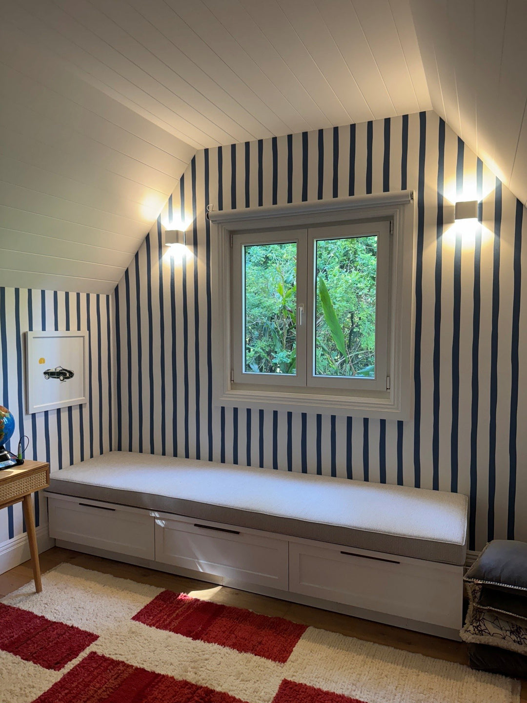 Built-in window seat beneath a window, with blue striped wallpaper and wall lights